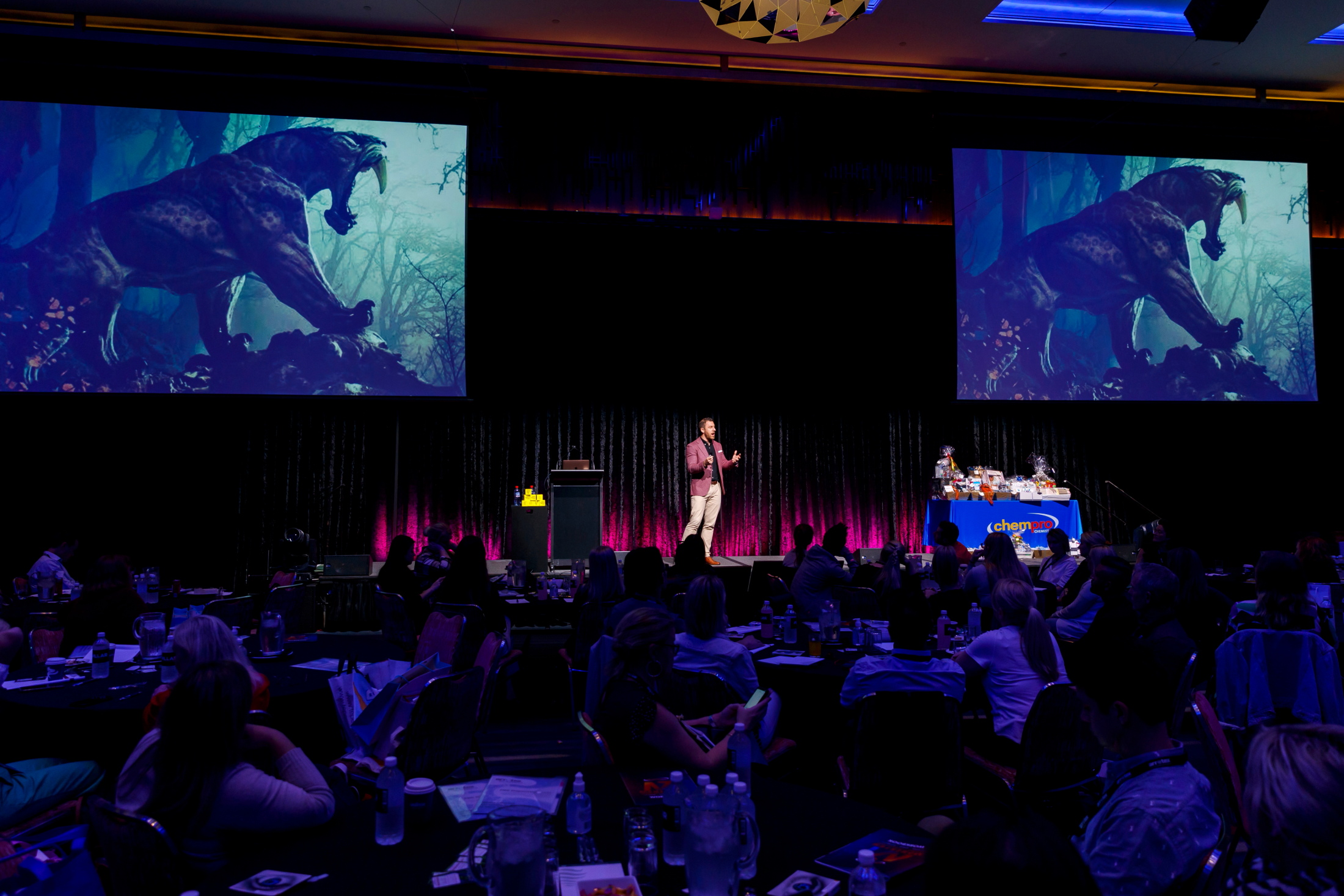 Olly Bridge speaking on a large conference stage with sabre-toothed tiger visuals on two large screens
